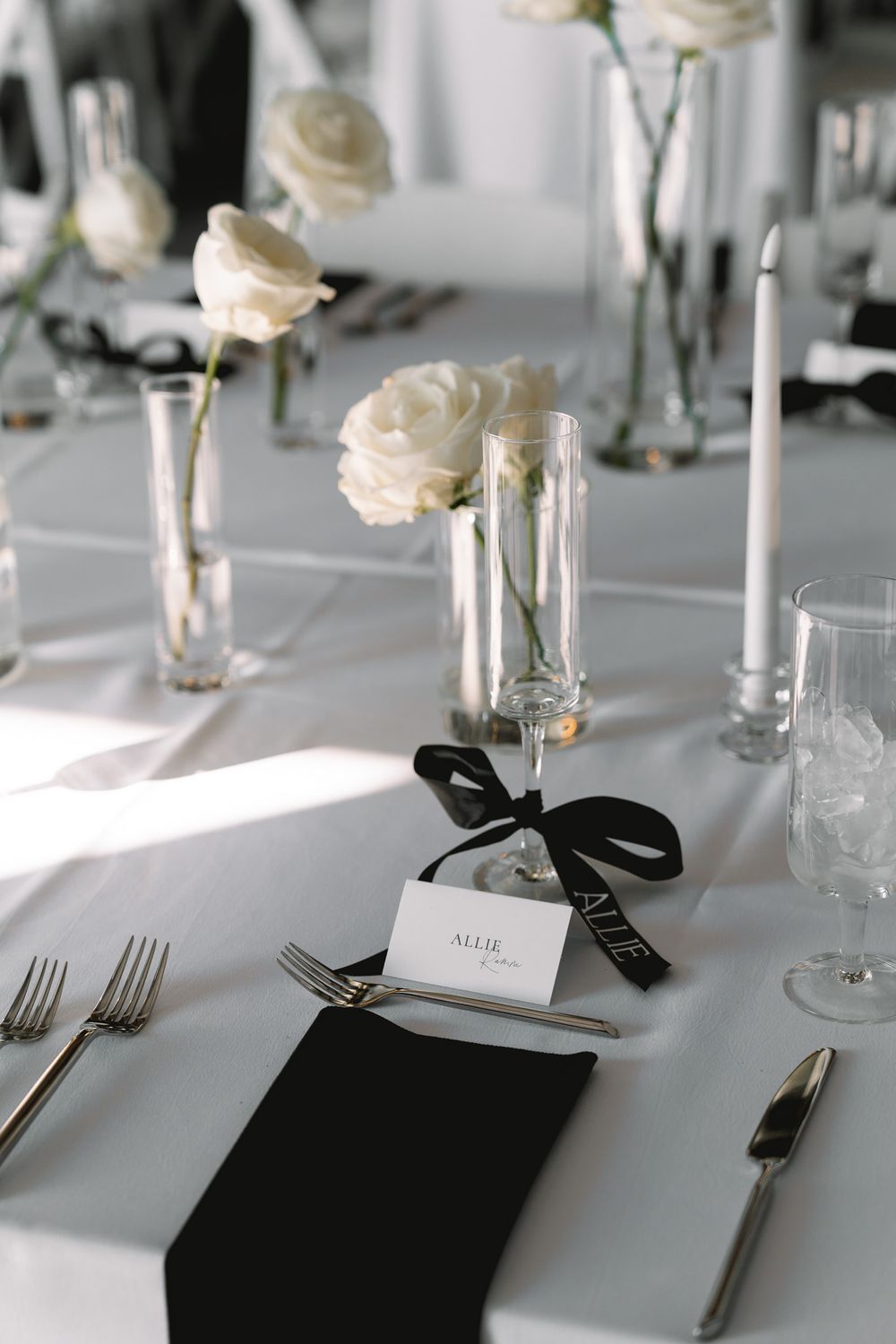 Elegant table setting with white roses in bud vases, black napkins, and place cards tied with black ribbon.