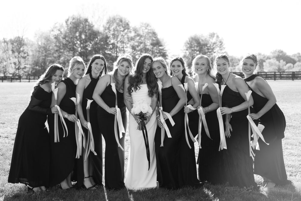 A wedding party portrait with bridesmaids in black dresses posing together with the bride in white on a sunny day.