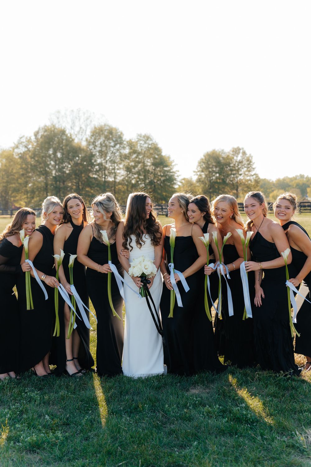 A bridal party wearing black bridesmaid dresses poses and laughs together with colorful ribbons during an outdoor wedding.