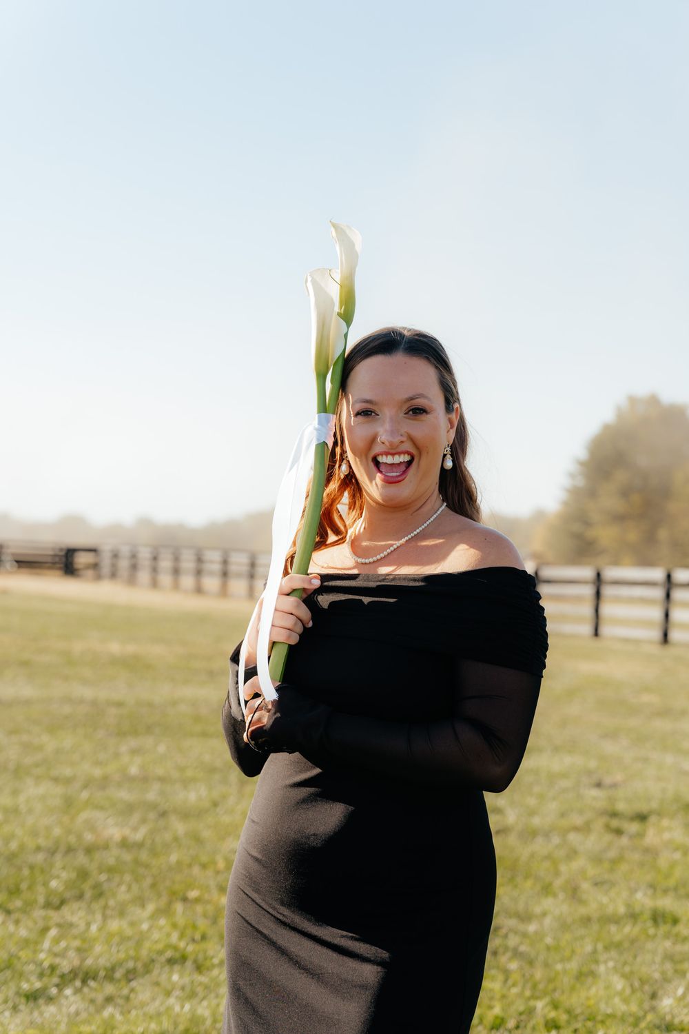 Someone in a black off-shoulder dress stands in a sunny field holding a calla lily flower.