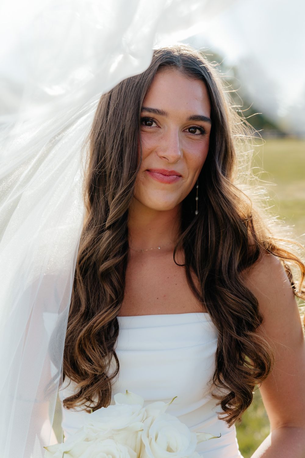 A bride in a white strapless wedding dress smiles while her long brunette hair glows in natural sunlight.