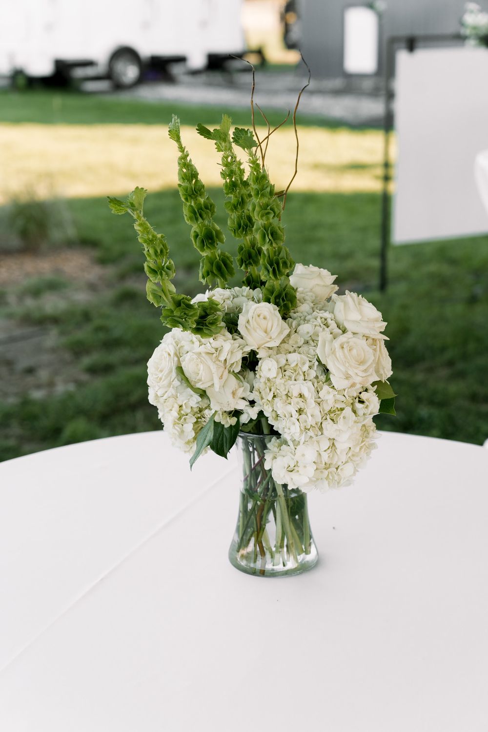 White hydrangeas and green bells of Ireland arranged in a clear glass vase on a white tablecloth at an outdoor event.