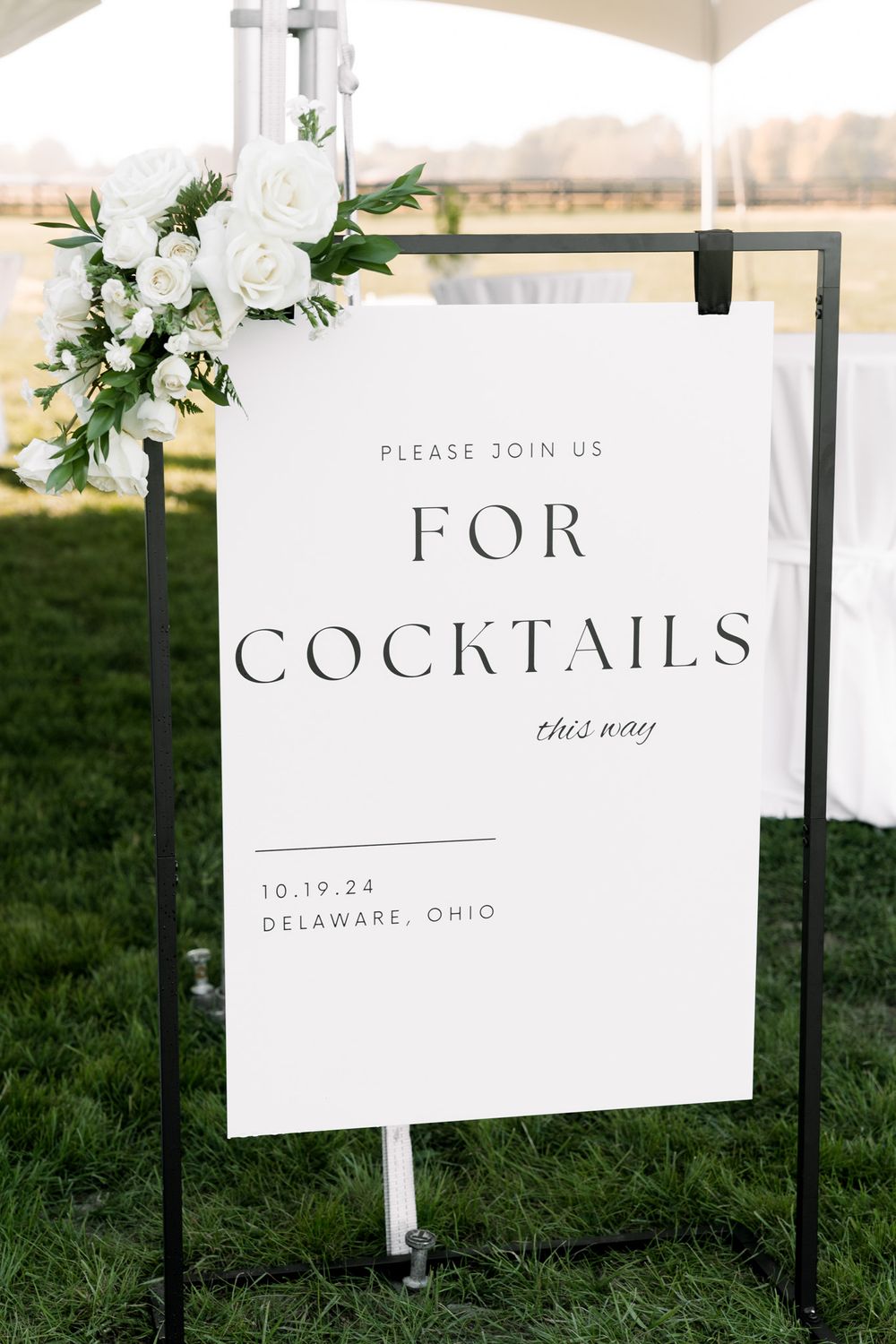 Elegant white cocktail sign with floral arrangement displayed on metal stand for outdoor wedding in Delaware Ohio.
