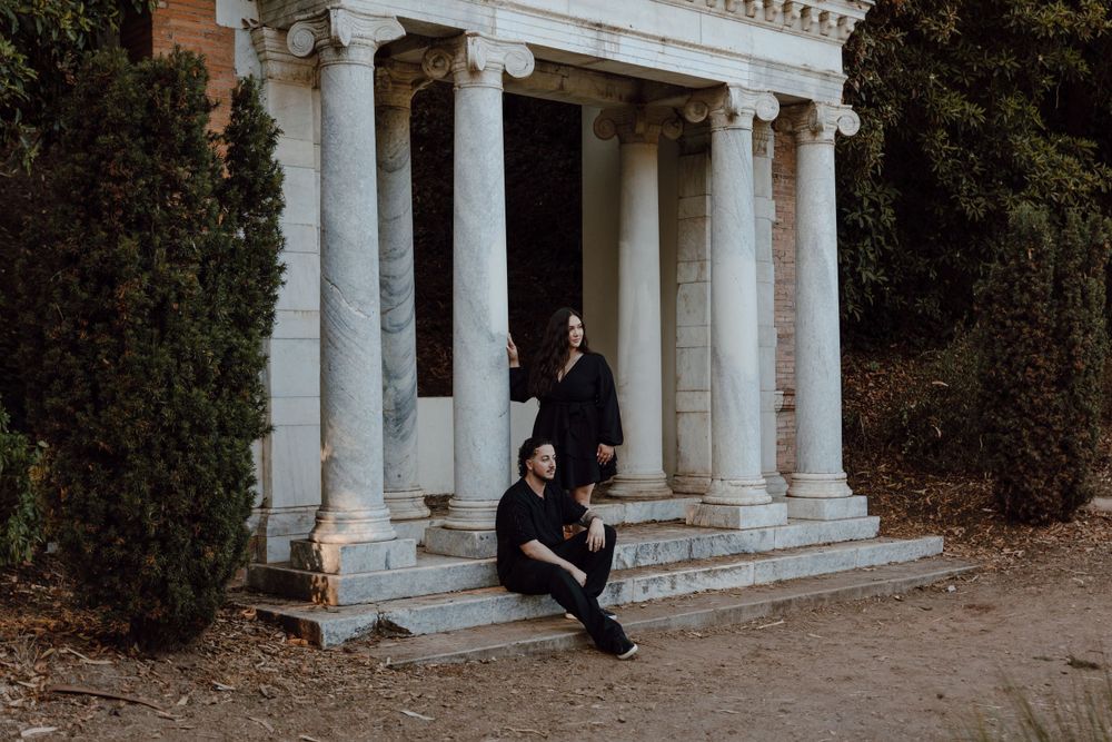 Two people sit on marble steps of a classical white columned structure surrounded by greenery at dusk.