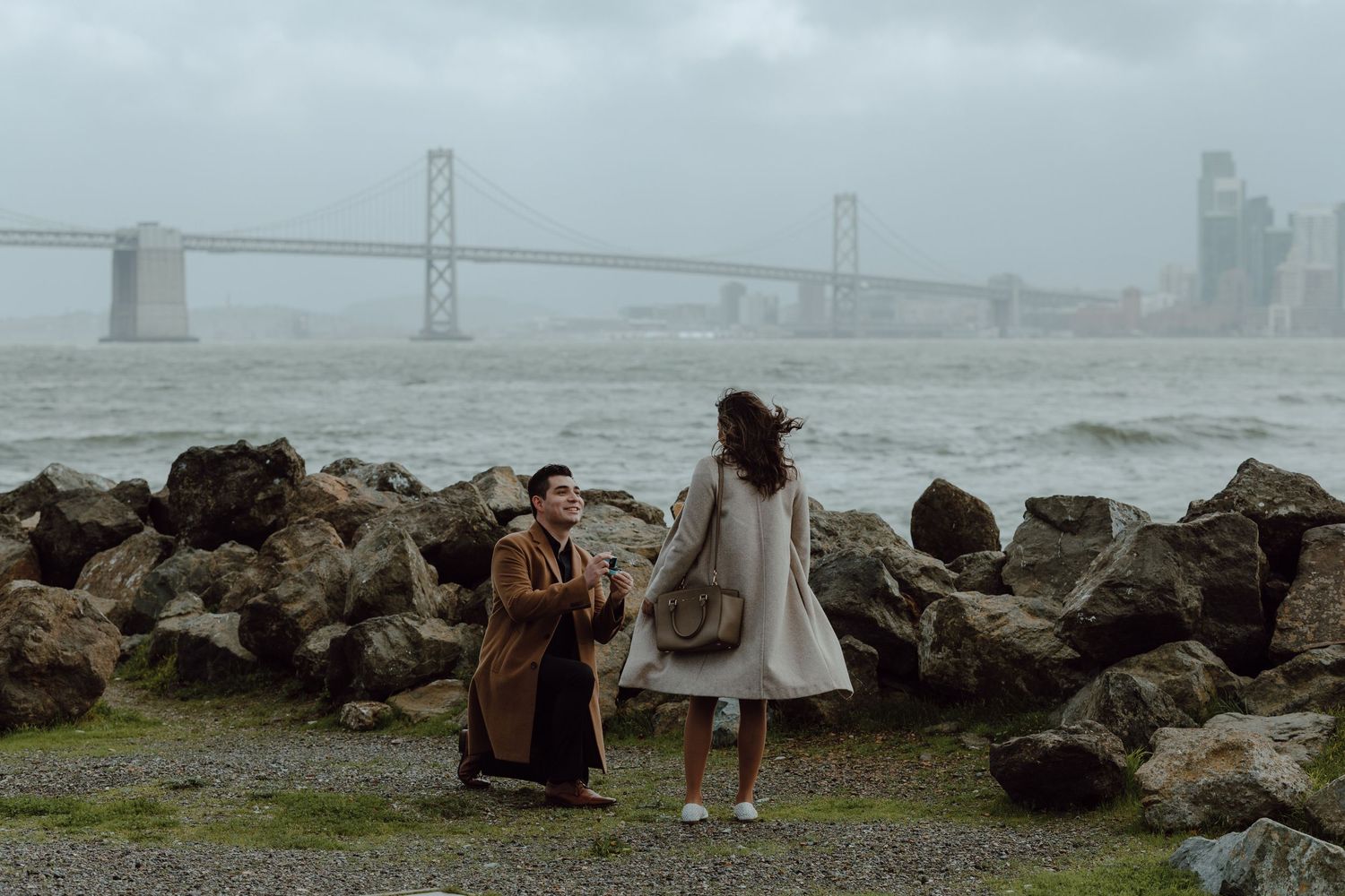 Couple stands on rocky shore gazing at San Francisco Bay Bridge on misty day.