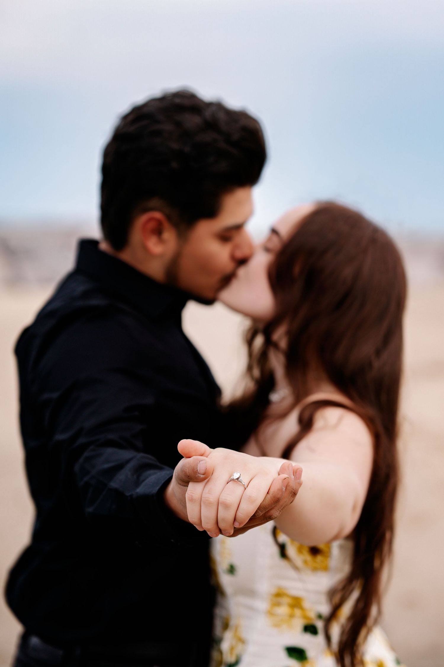 A romantic couple shares a tender kiss while holding hands in a dreamy desert landscape with soft lighting.