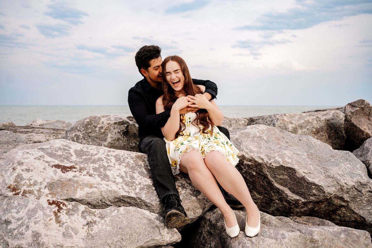 A couple shares a playful moment while sitting together on rocky coastal terrain under cloudy skies.