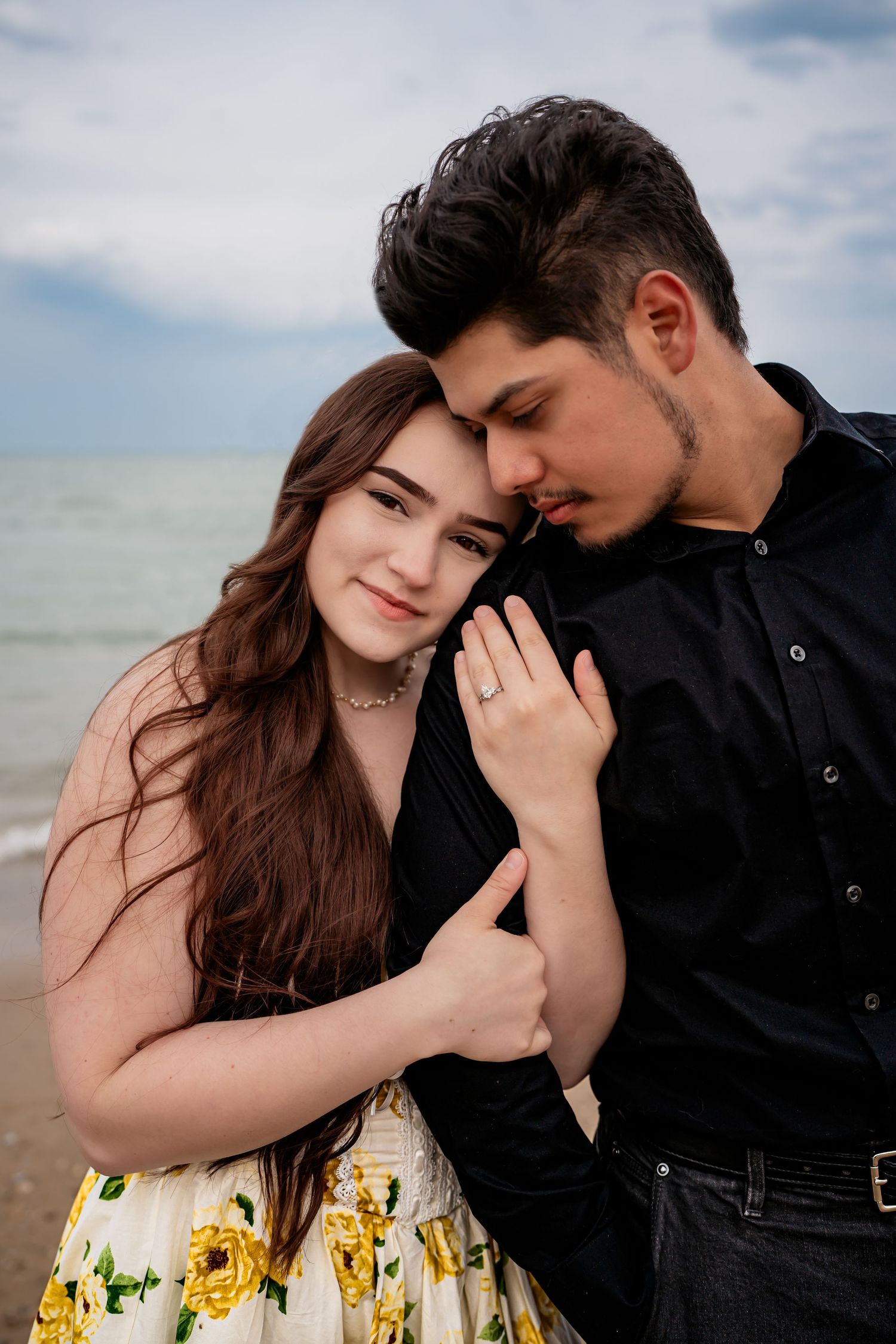 A romantic couple embraces on a beach, with her wearing a floral dress and him in dark clothing as waves roll in behind them.