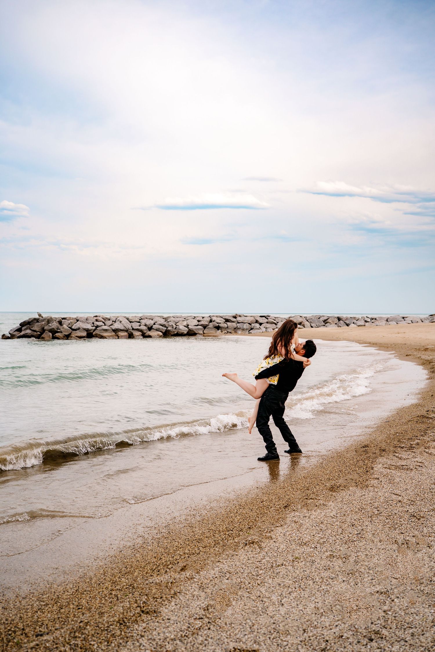 A romantic seaside moment with two dark-clothed figures embracing on a sandy beach near a rocky jetty at sunset.