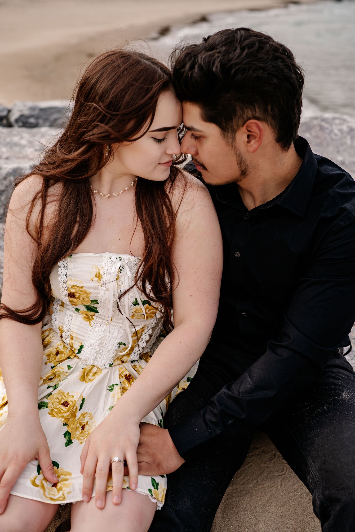 Romantic couple sitting close together outdoors, her in a floral sundress and him in black attire.