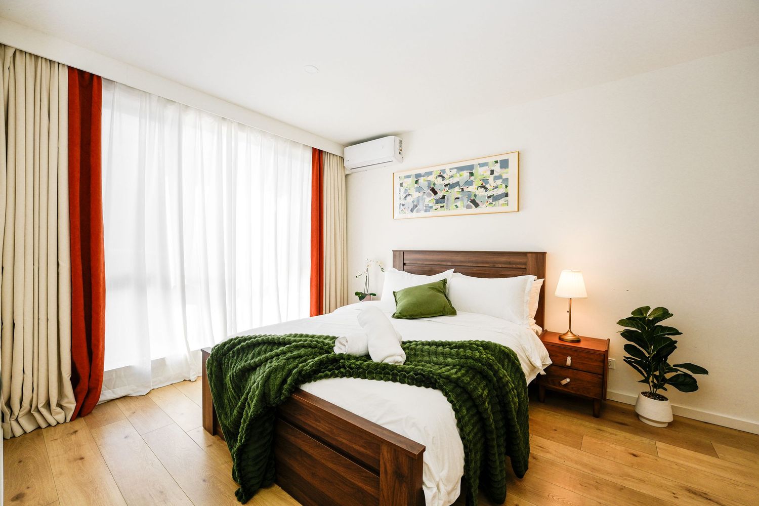 Modern bedroom with wooden bed frame, white bedding, green throw blanket, orange curtains and houseplant in corner.