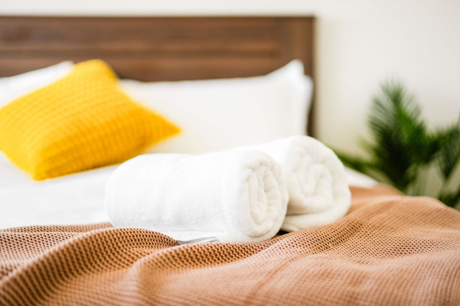 Cozy bed with yellow accent pillow, white rolled towels, and brown knitted blanket beside green plant decor.