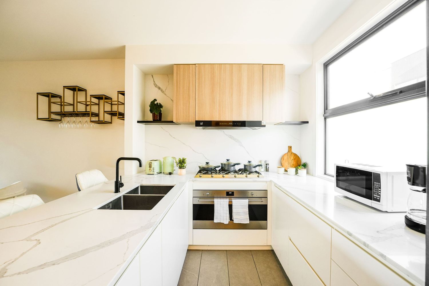 Modern white kitchen with marble countertops, wooden accents, and large window featuring minimalist design elements.