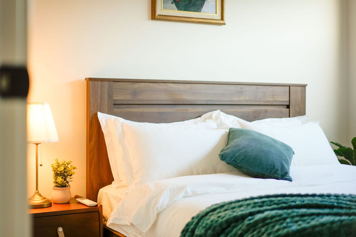 Cozy bedroom interior with wooden headboard, white bedding, teal accent pillow, green throw blanket and bedside lamp.