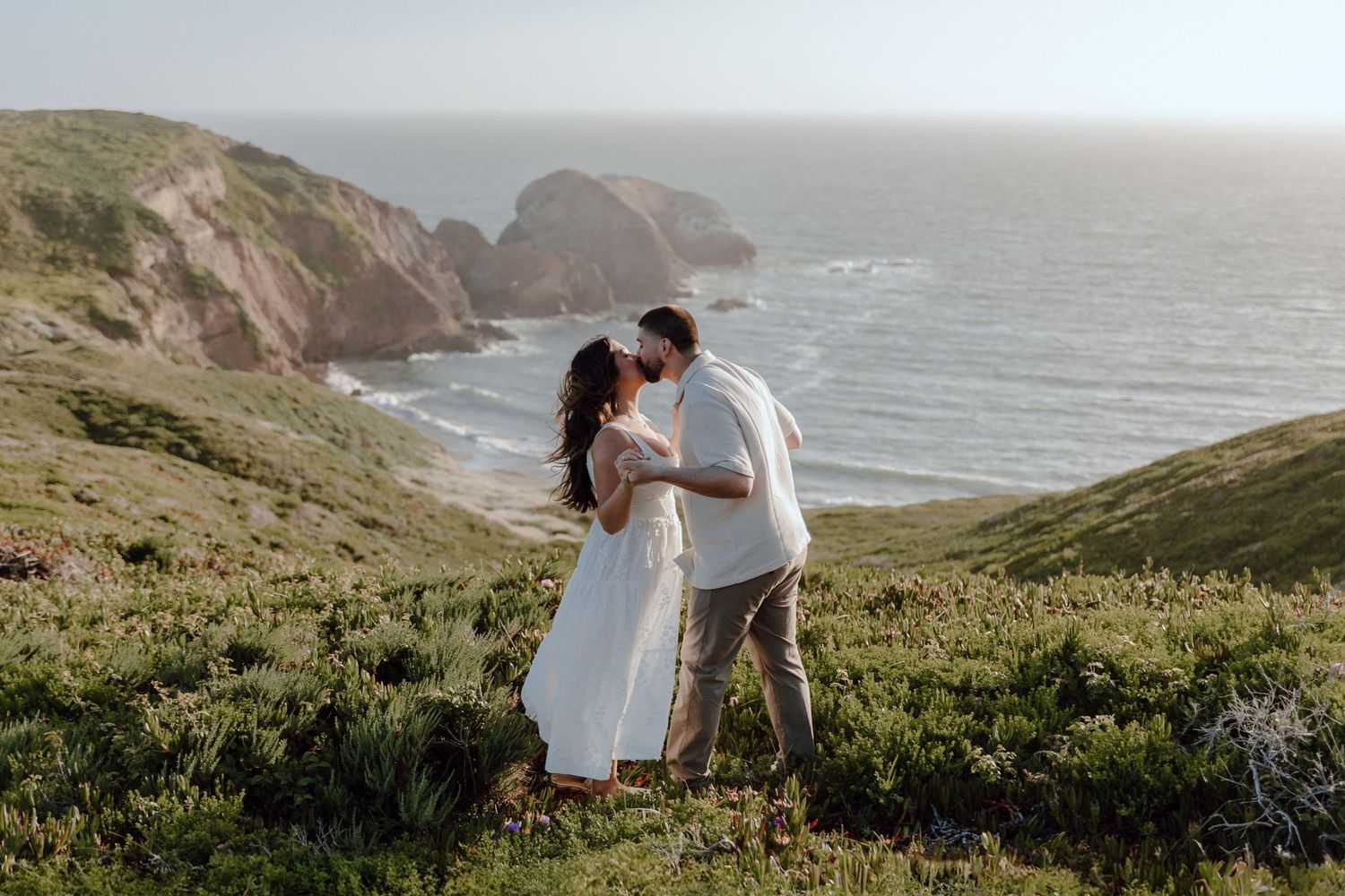 A romantic couple shares a tender moment on a pathway through golden grass at sunset in a coastal setting.