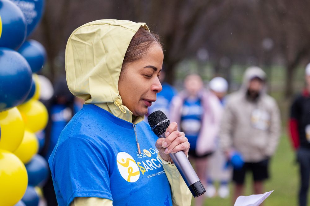 A speaker in a yellow hood and blue shirt addresses a crowd at an outdoor event with blue and yellow balloons.