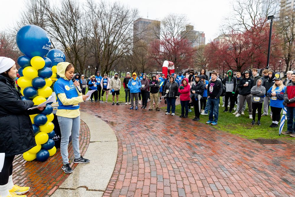 Crowd gathers at outdoor event with blue and yellow balloon decorations on brick pathway near bare winter trees.