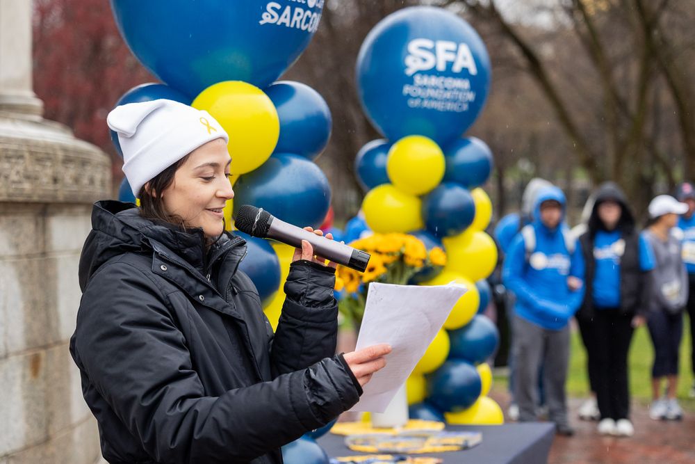 Student in winter coat speaks into microphone at outdoor event with blue and yellow balloon decorations in background.
