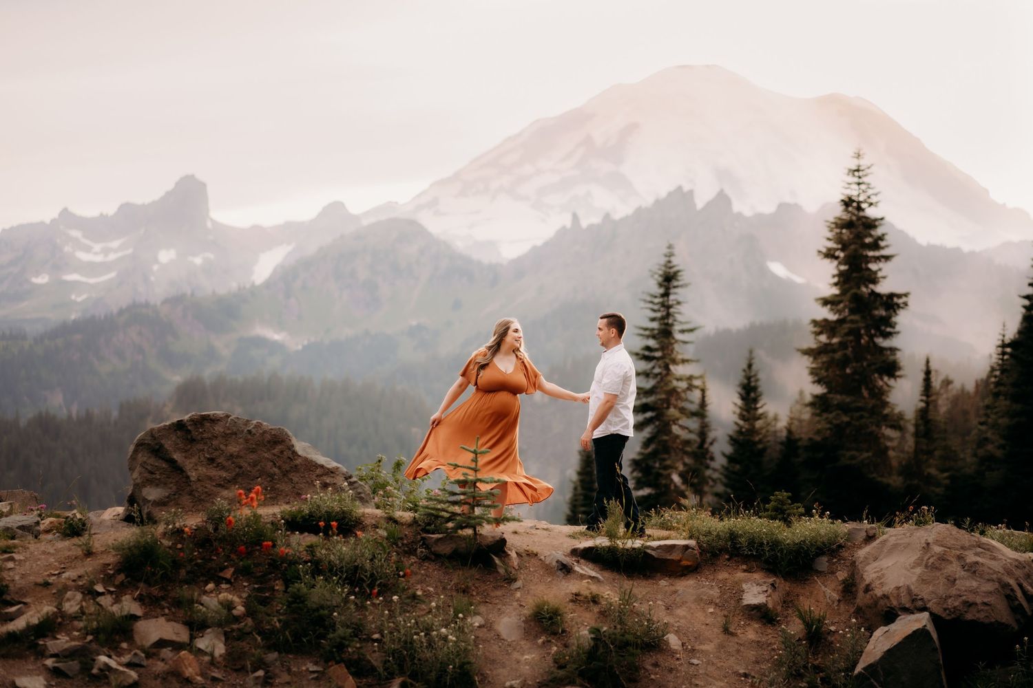 Pregnant woman in orange dress dances with her husband atop a mountain side with tall trees and Mt. Rainier in the background.