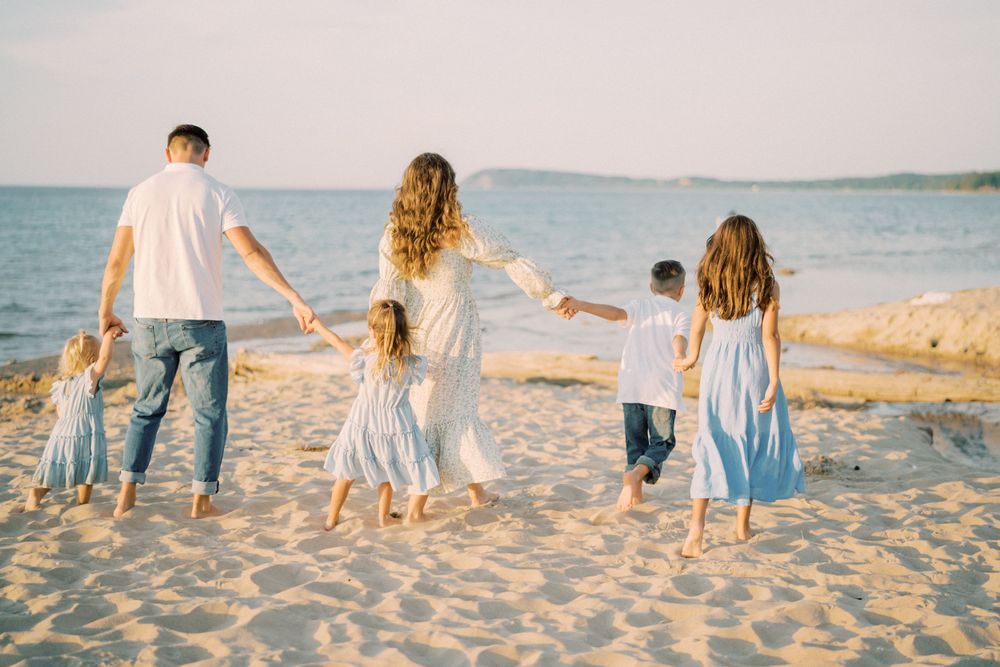 Family holding hands while walking together on beach sand near ocean at sunset.