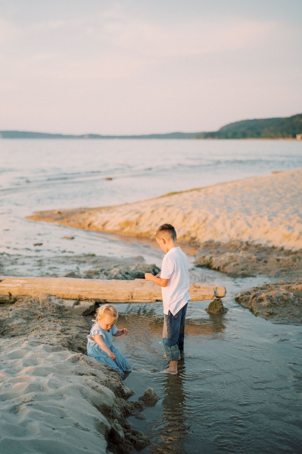 Two figures play in the shallow water at the beach during sunset with hills in the background.