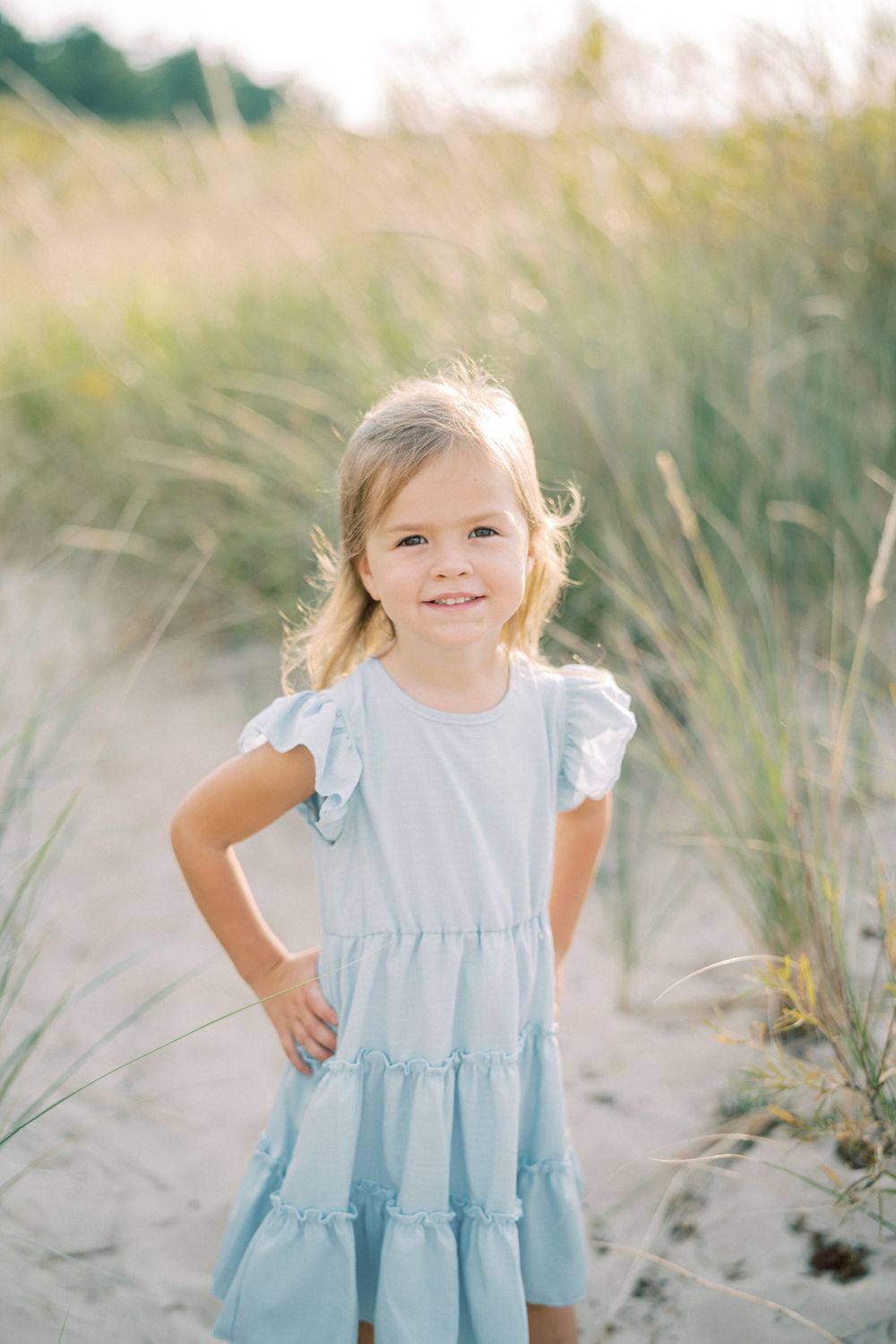 Young child in light blue ruffled dress posing confidently among beach grass.