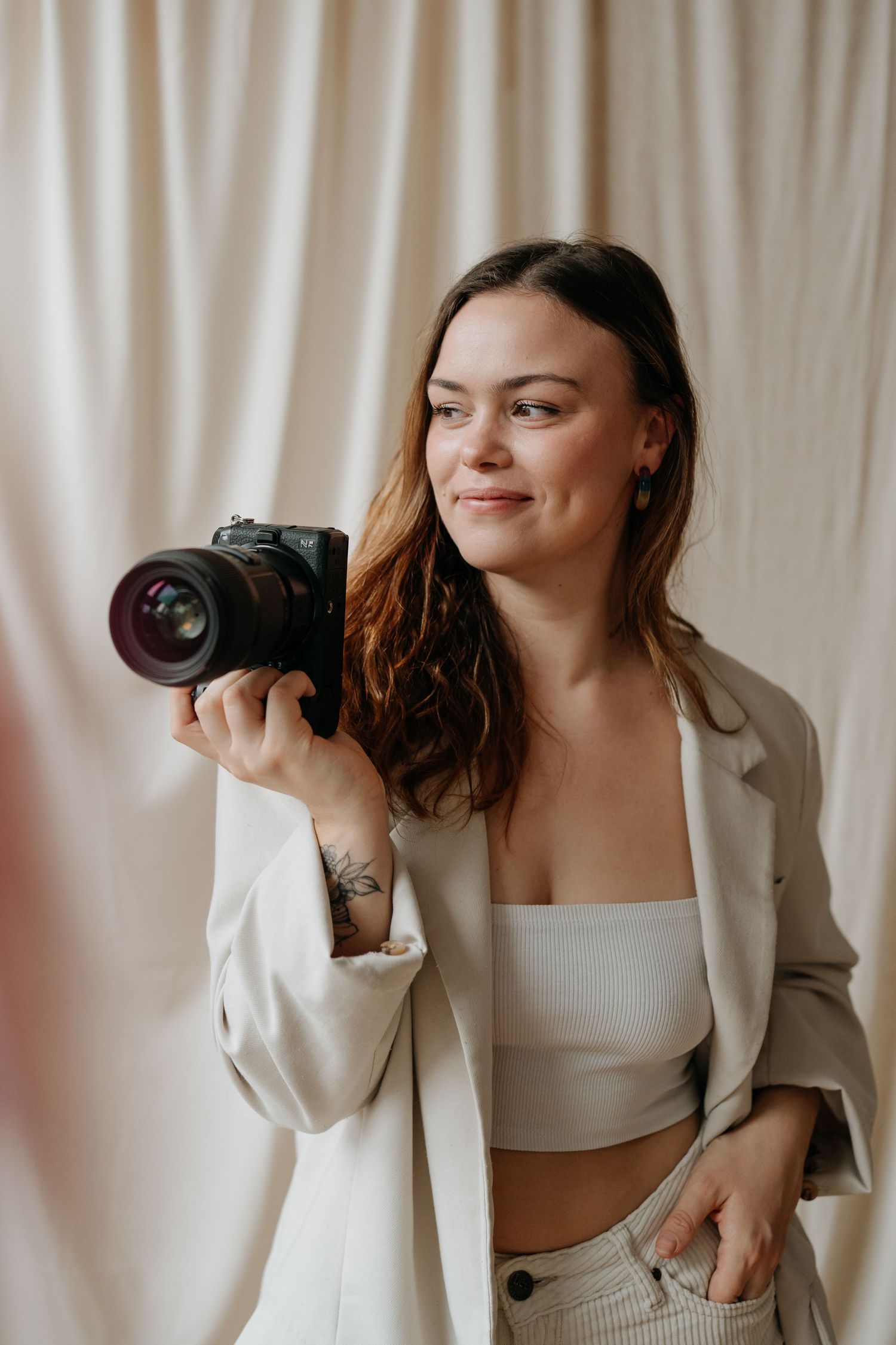 Professional photographer in white blazer and crop top holding DSLR camera against light backdrop.