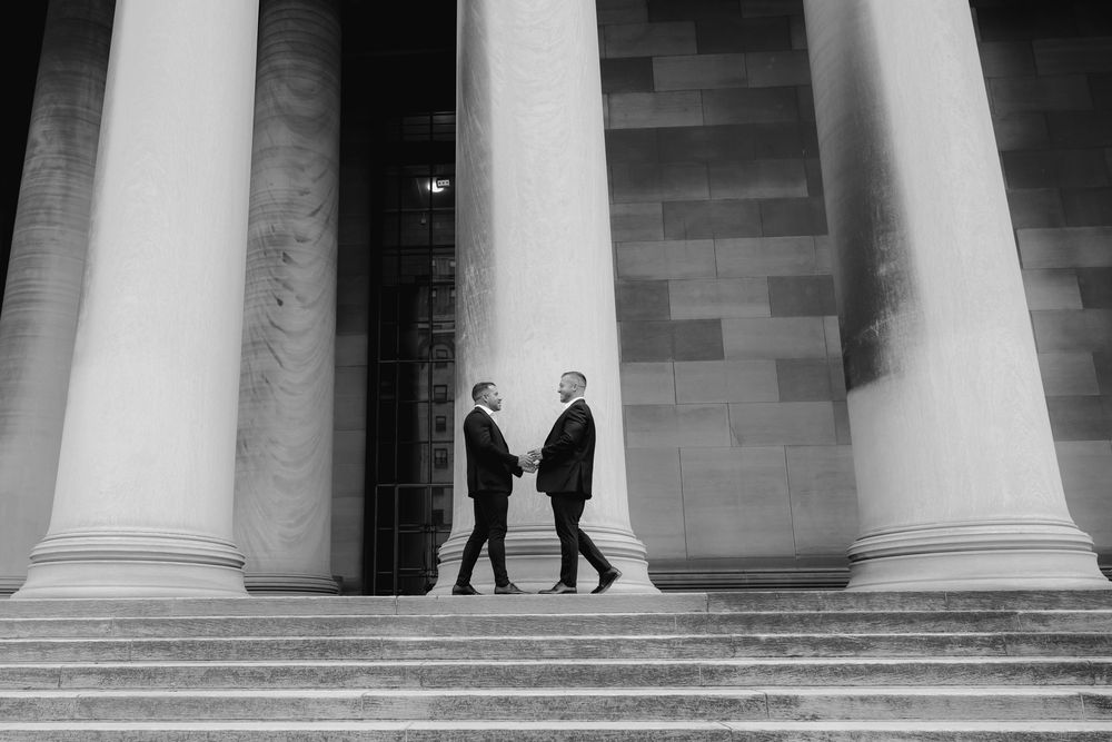 Two figures stand between massive white columns on stone steps in a dramatic black and white architectural composition.