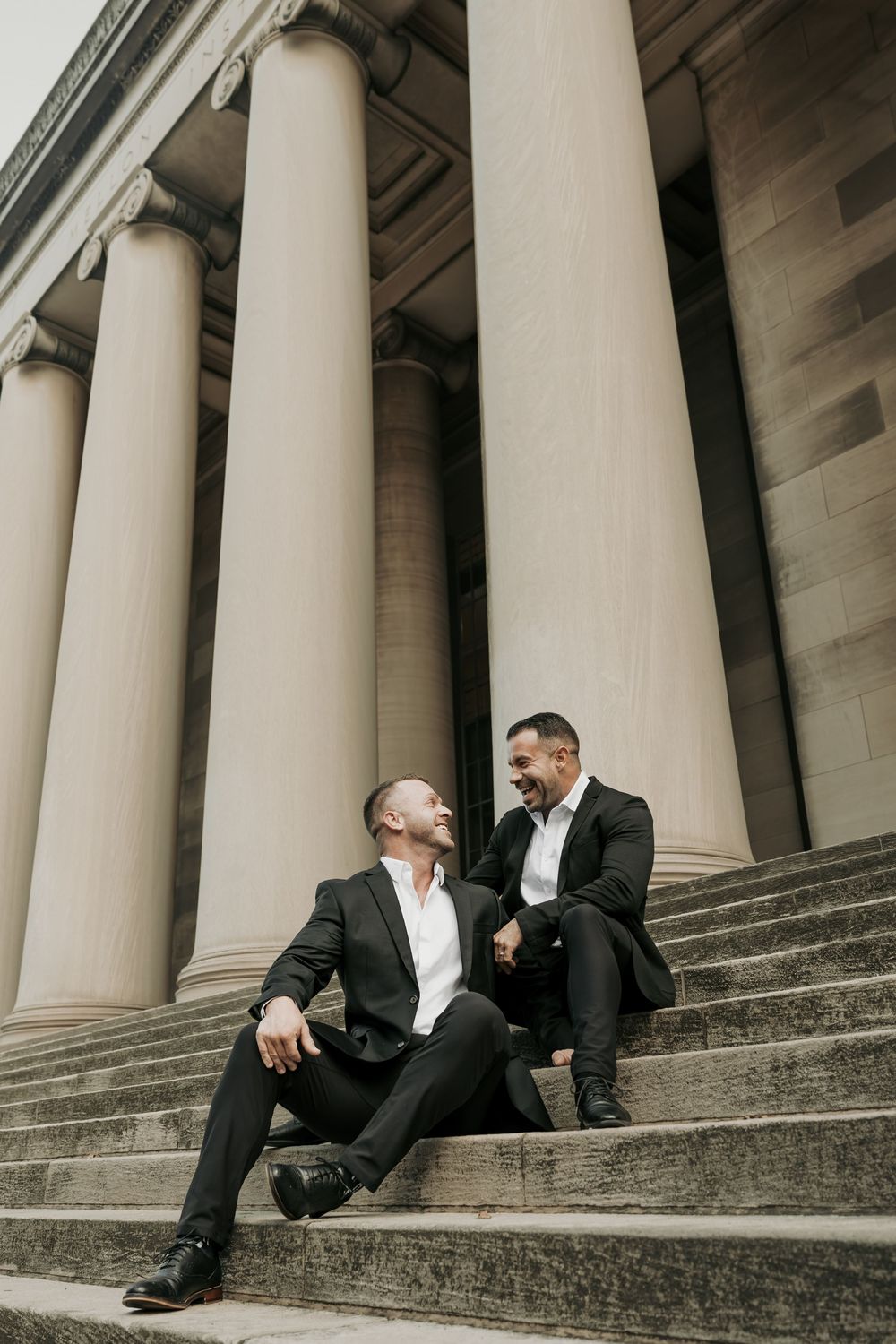 Two people in formal black suits sit on grand stone steps between towering neoclassical columns.