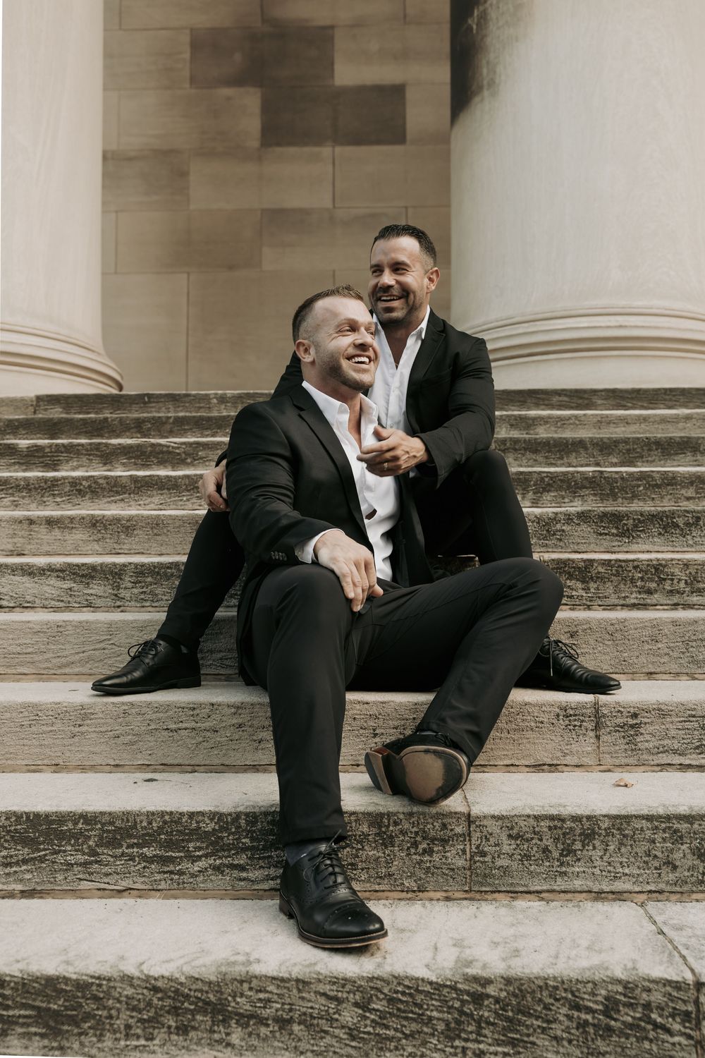 Two people in black suits sitting and laughing together on stone steps of a building.