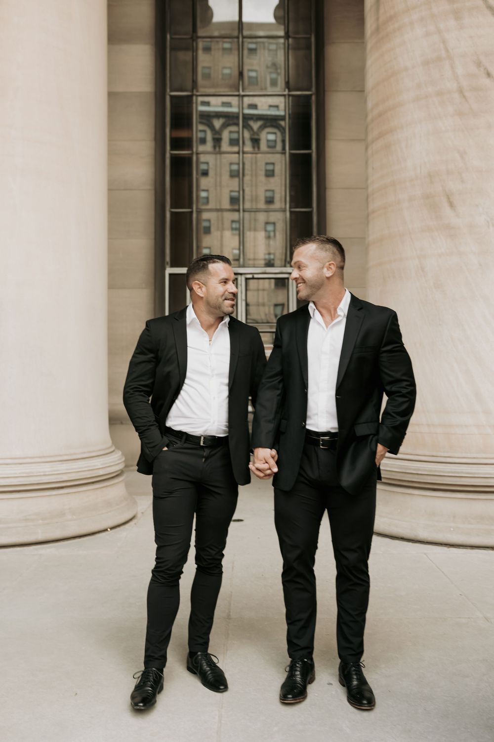 Two people in matching black suits and white shirts stand holding hands between elegant stone columns.