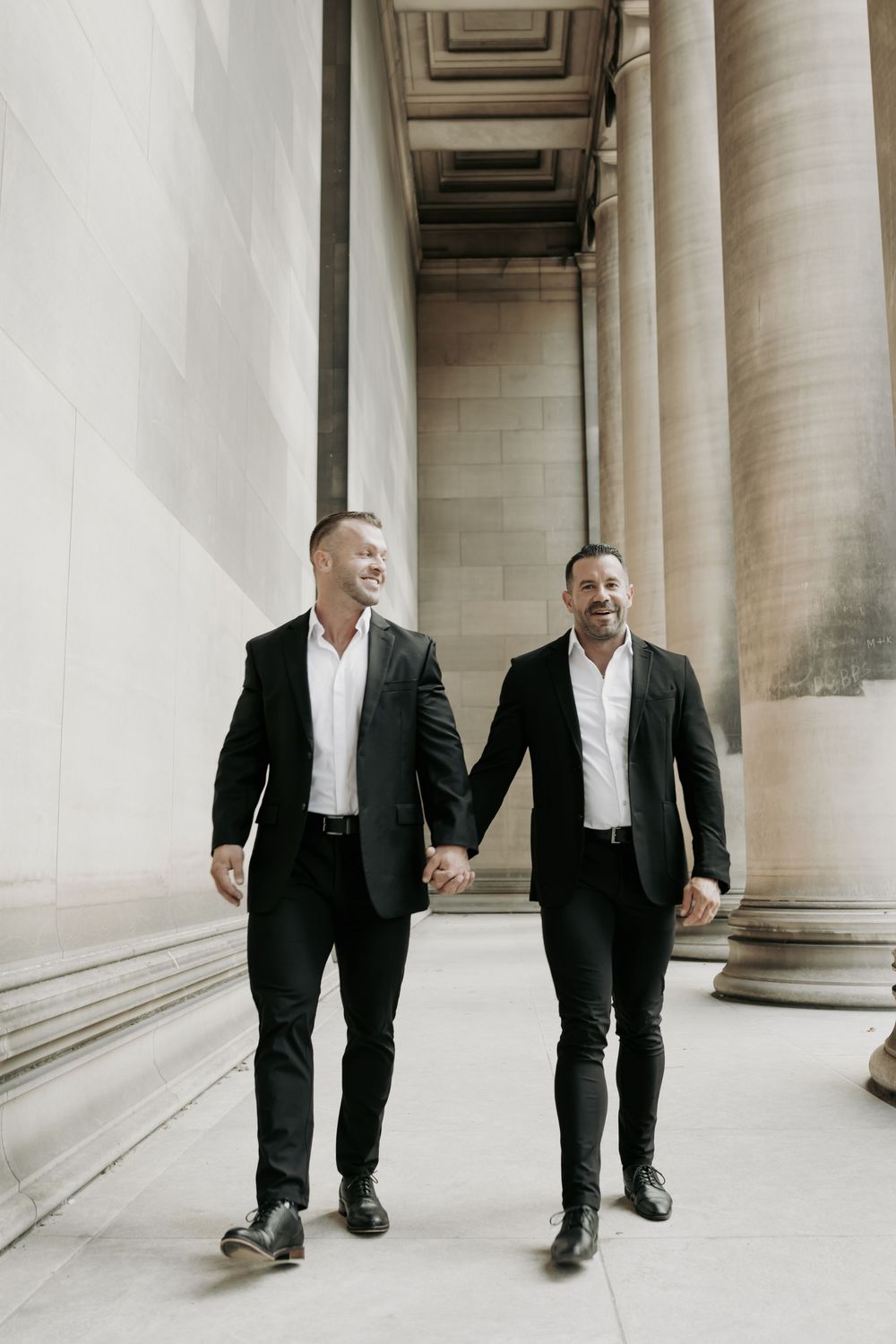 Two people in matching black suits and white shirts walk hand in hand between elegant stone columns of a grand building.