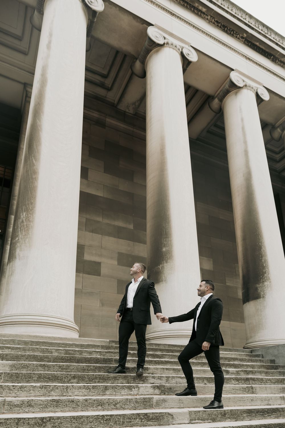 Two figures in black suits stand on grand stone steps between towering classical white columns of a historic building.