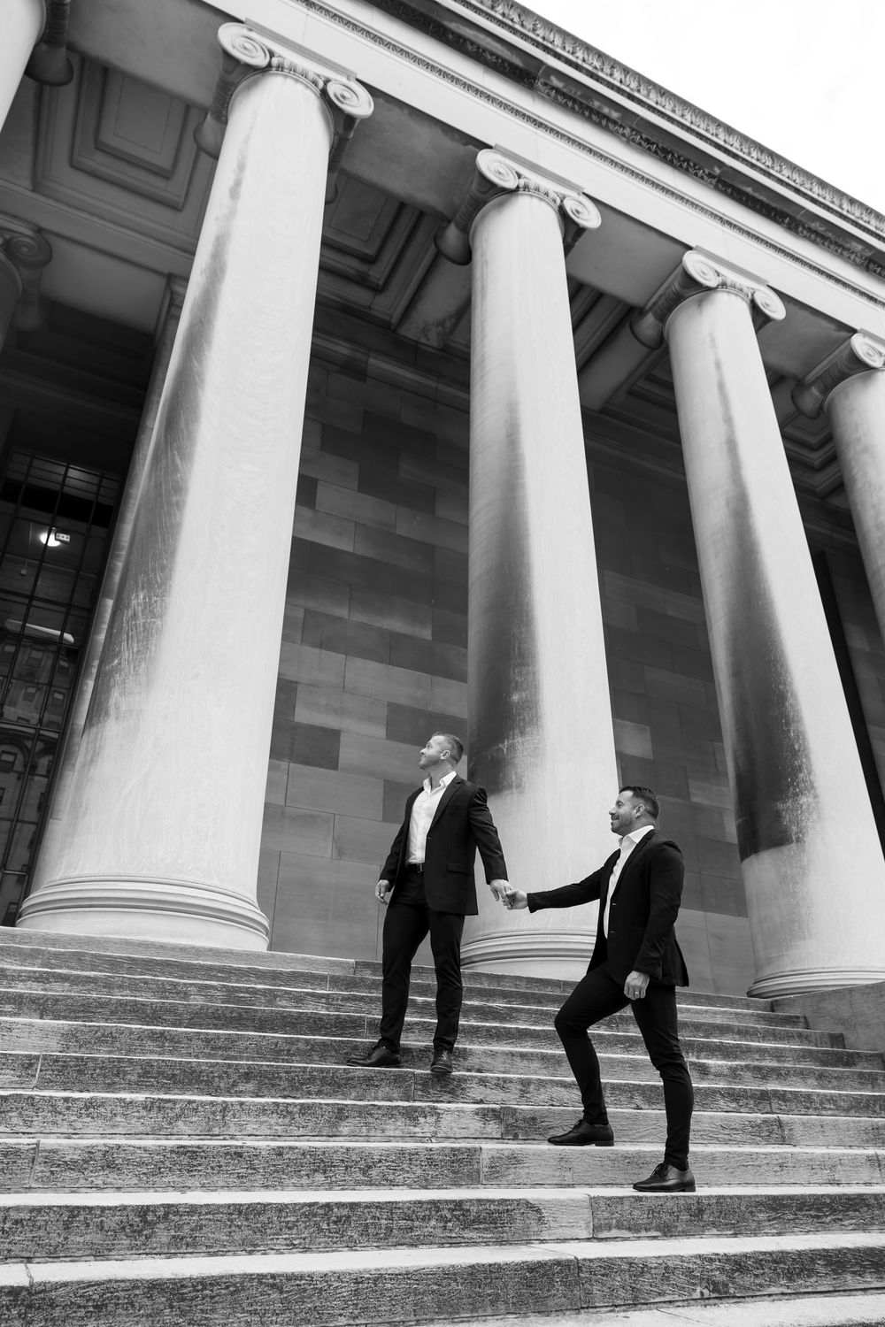 Two people in formal black attire ascending grand stone steps with tall classical columns in the background.