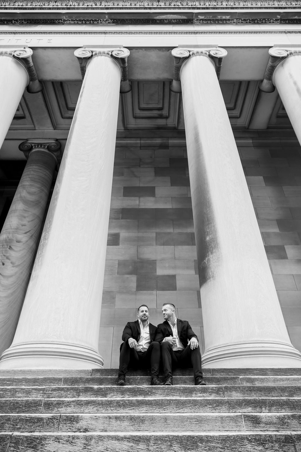 A black and white architectural photograph showing two people sitting on steps between grand classical columns.