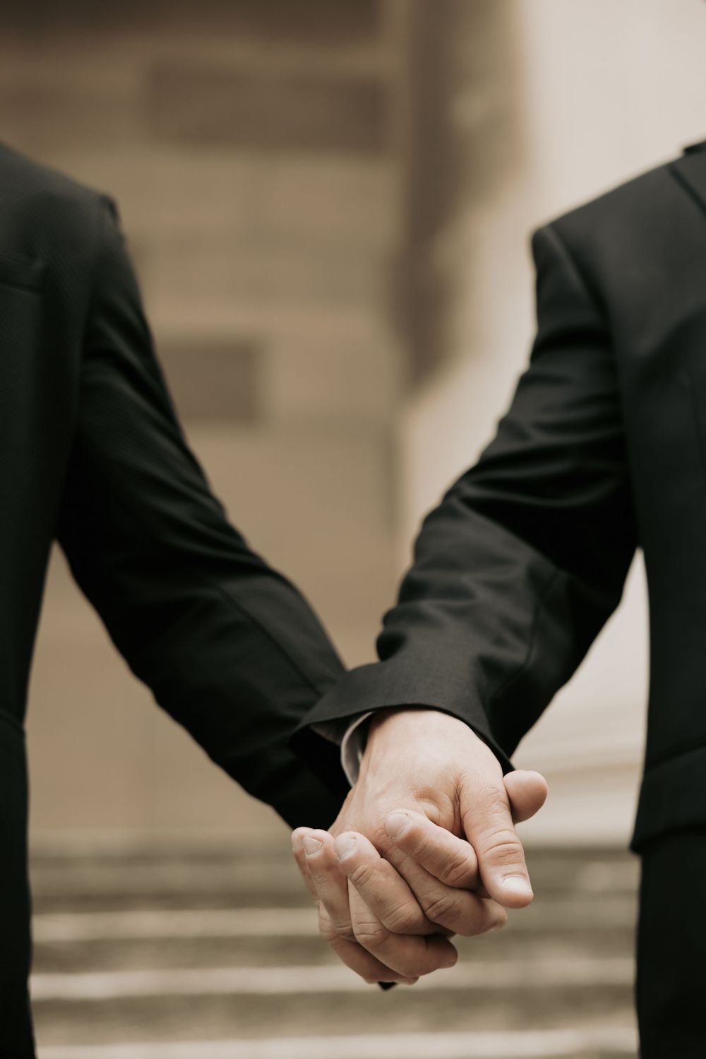 Close-up shot of two people in dark suits holding hands while standing on stone steps.
