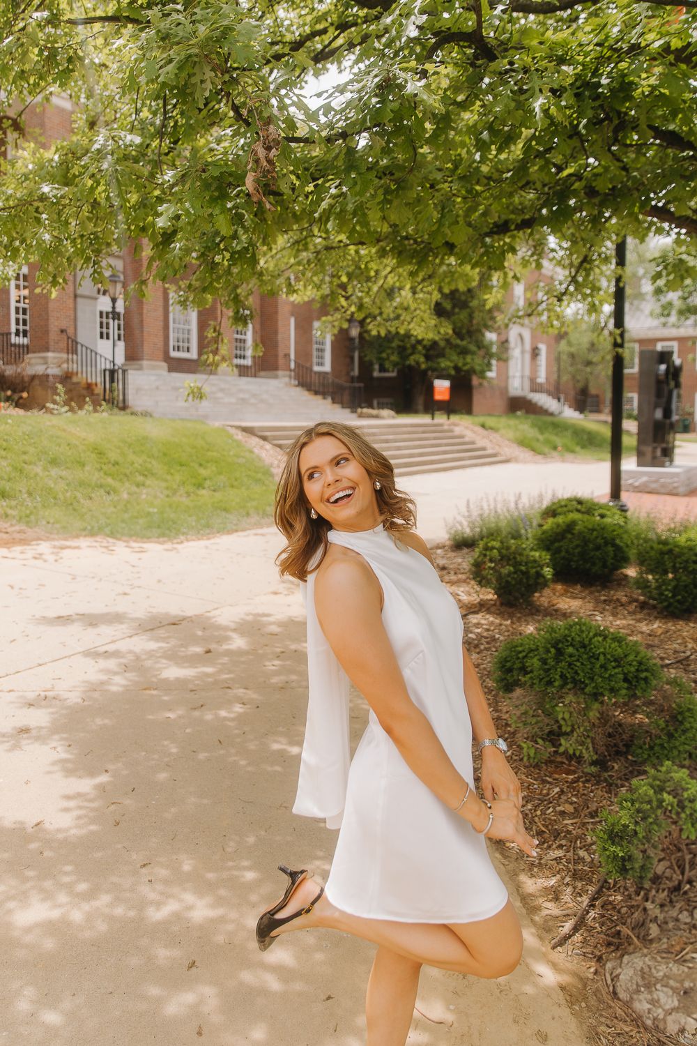 Someone in a white sleeveless dress twirls playfully on a sunny path near brick buildings and green landscaping.