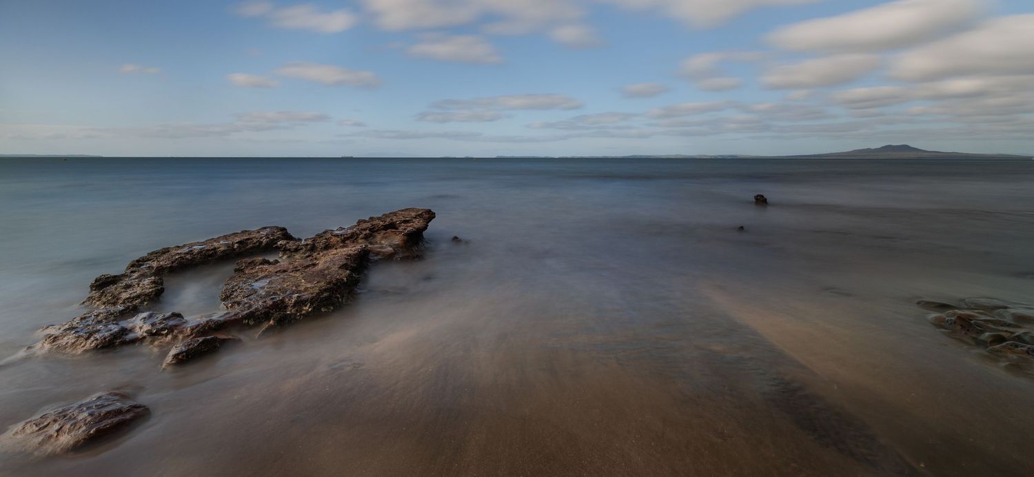 Rugged rocky outcrop emerges from smooth beach waters under cloudy skies with distant island on horizon.