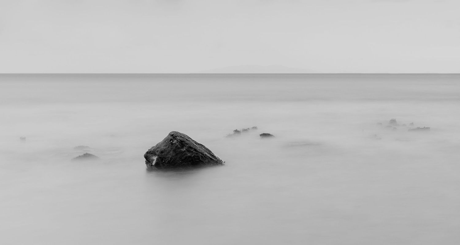 A dark rock formation emerges from misty waters in a minimalist black and white seascape.