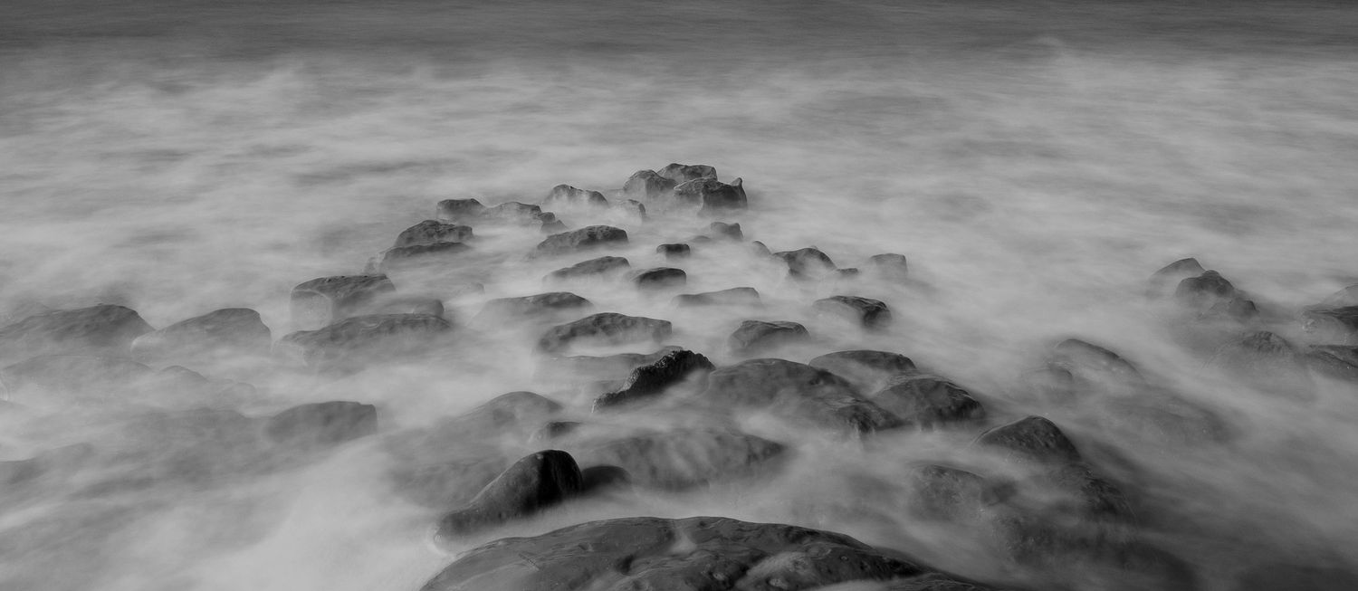 Long exposure black and white seascape shows misty waves flowing over rocky shore in ethereal motion.