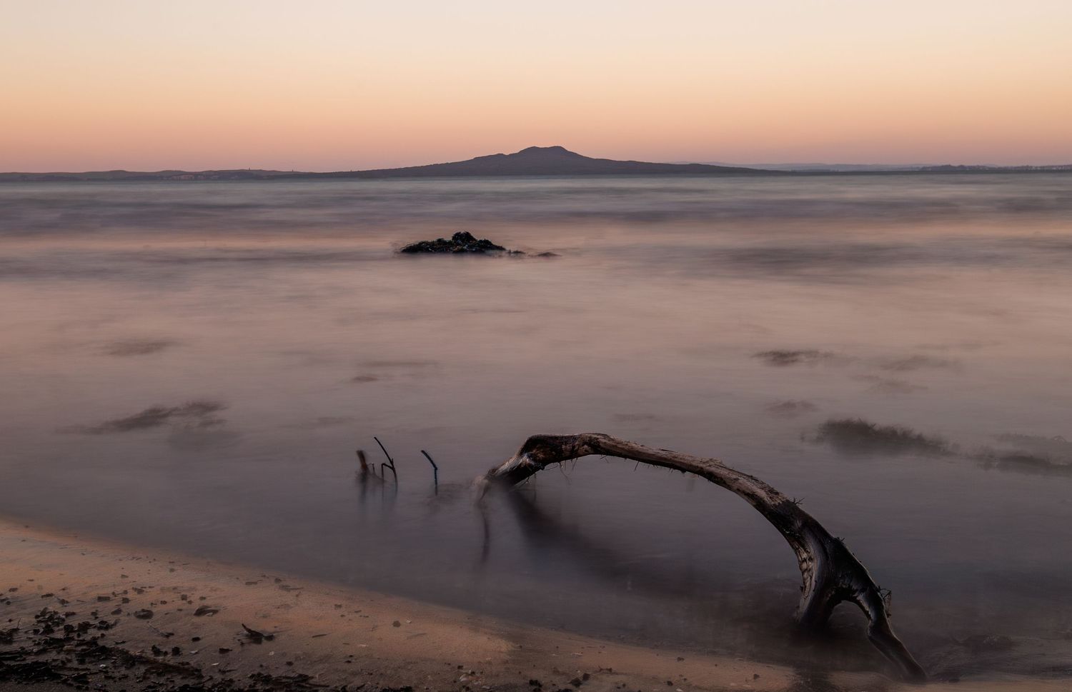 Misty seascape at sunset with driftwood on beach and distant mountain silhouette on the horizon.