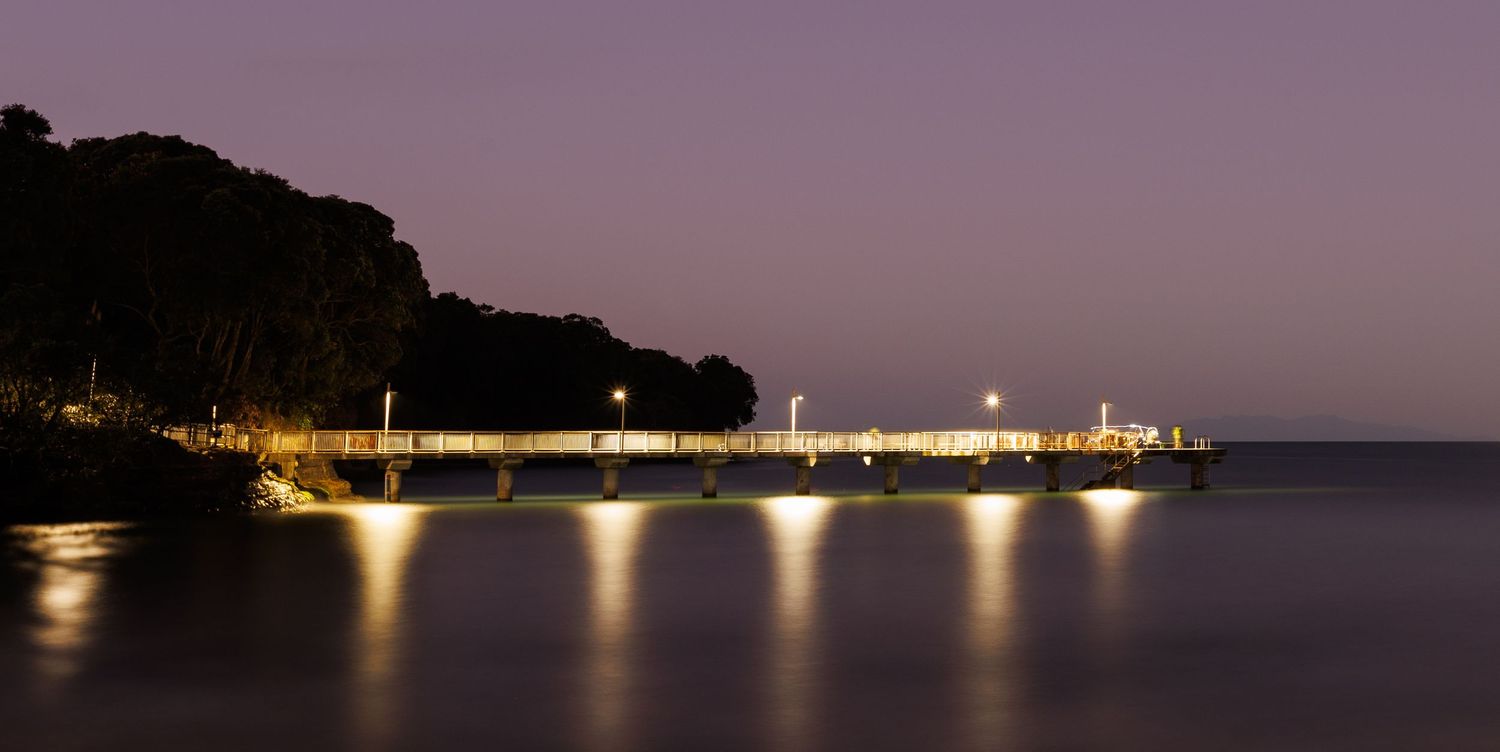 Illuminated waterfront pier glows against purple twilight sky with reflections dancing on the dark water.
