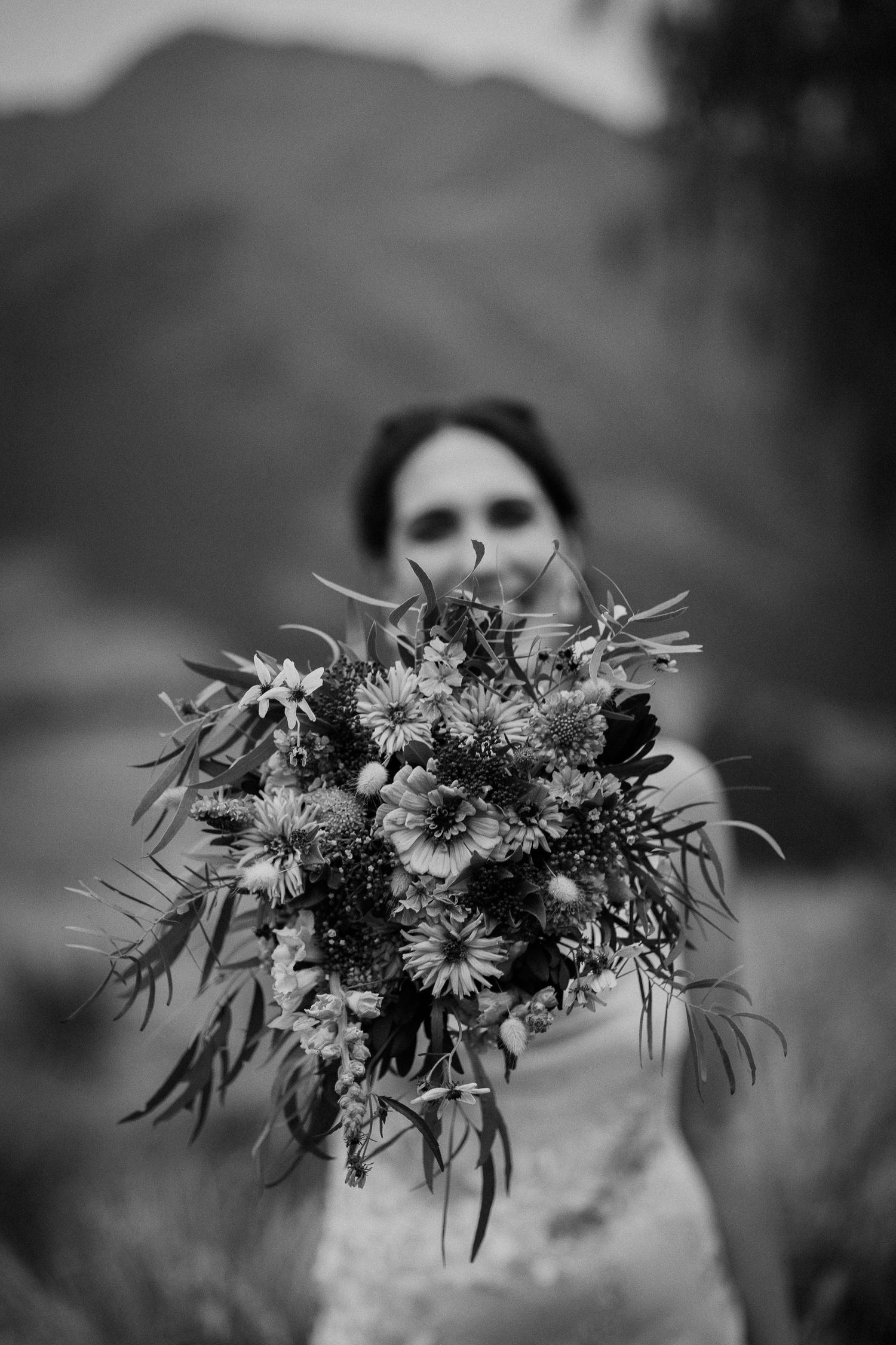 A black and white photograph showing a rustic wildflower bouquet held up against a blurred mountain landscape.