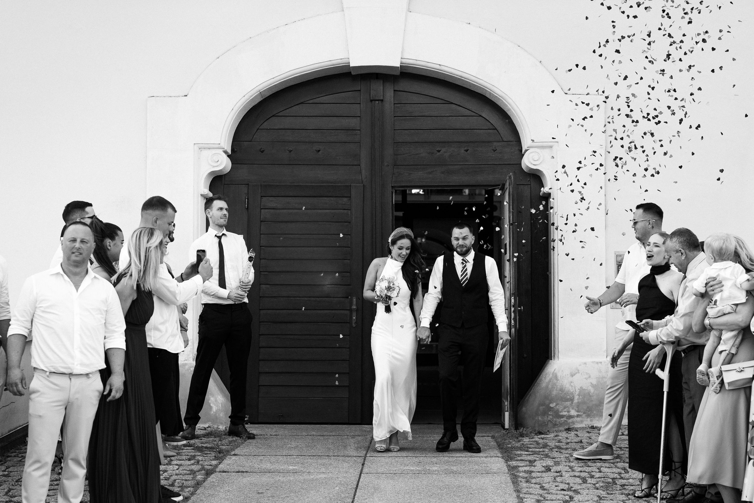 A black and white photograph of a wedding couple exiting through an arched doorway while guests shower them with confetti.