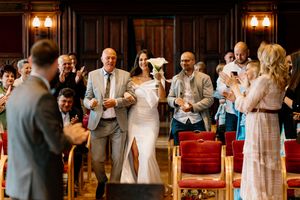 Wedding guests gather in an ornate wood-paneled venue with warm lighting and red seating during a ceremony.