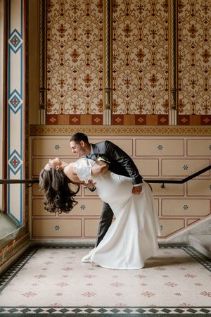 Elegant couple poses in a romantic dance dip against vintage wallpapered wall with decorative trim and stained glass window.