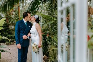 A romantic wedding moment captured in a greenhouse setting surrounded by lush tropical plants and white columns.