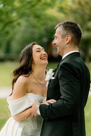 A joyful wedding couple shares a candid laughing moment together outdoors in a garden setting.