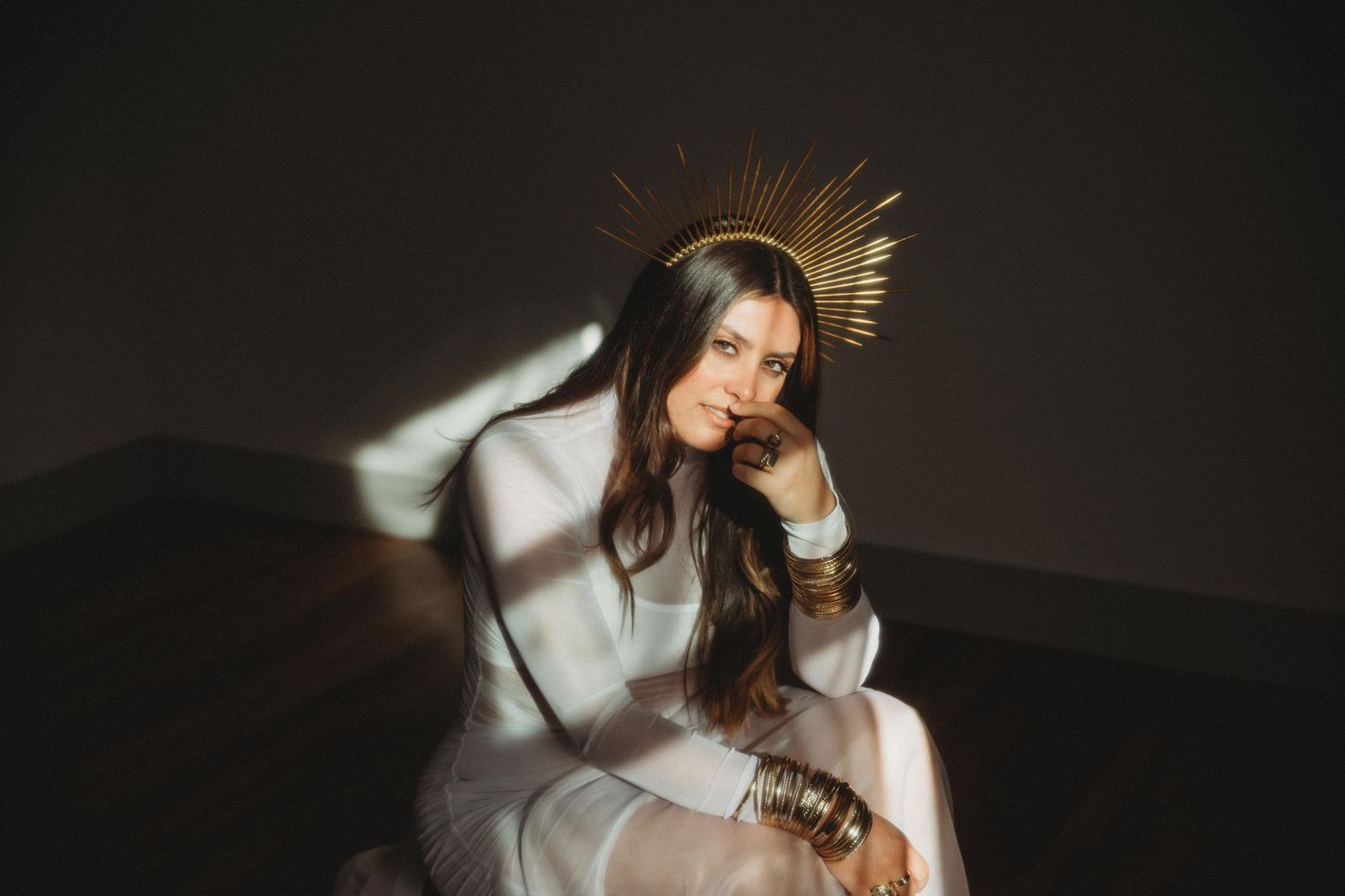 A person in a white dress sits thoughtfully in a darkened room with a dramatic light beam and crown-like headpiece.
