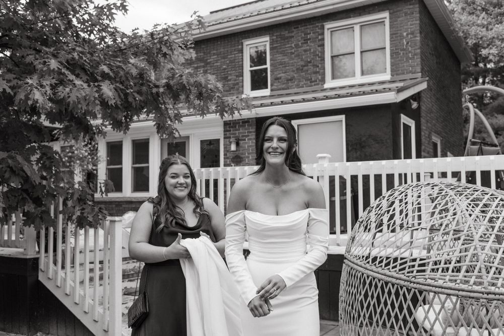 Two people in celebratory attire stand outside a brick house with a white picket fence in a black and white photo.
